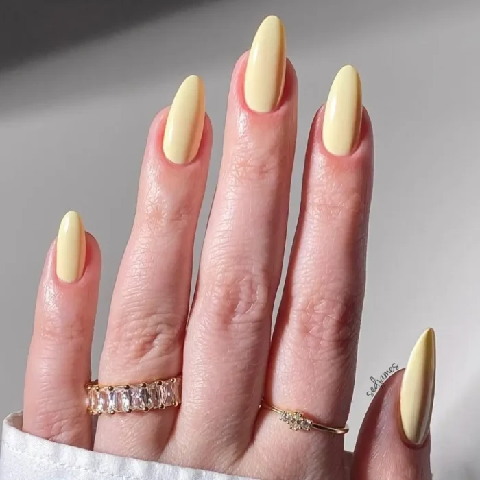 A hand with elegant, long almond nails showcasing a flawless manicure in a creamy butter-yellow polish.