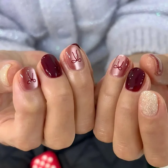 Short nails with deep berry and iridescent pink gel polishes, featuring delicate bow designs on some nails and glitter on the pinky.