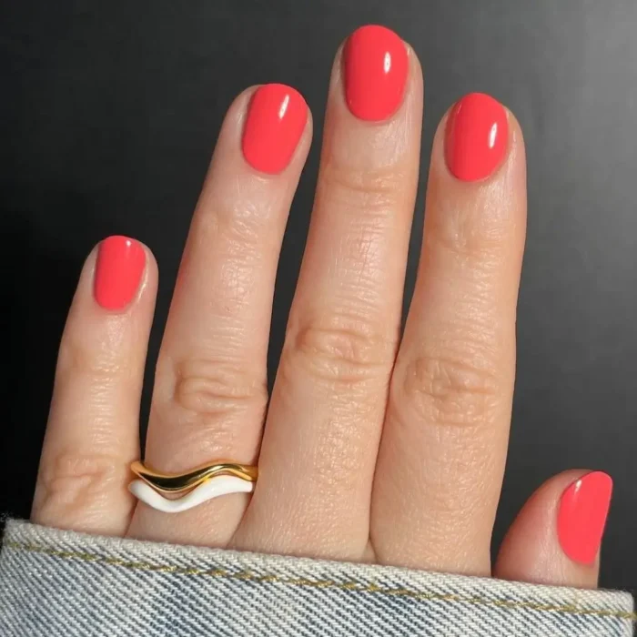 A close-up of a hand with a glossy, vibrant coral-pink manicure on short, rounded nails against a dark background.