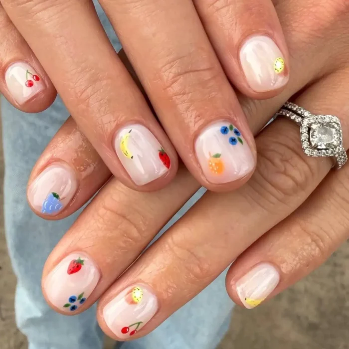Hands showing a minimalist fruit nail design on short natural nails, featuring a tiny mixed fruit pattern on a milky white base.