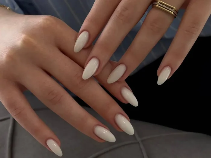A pair of hands showcasing a sophisticated milky white manicure on long, almond-shaped nails, adorned with gold rings.