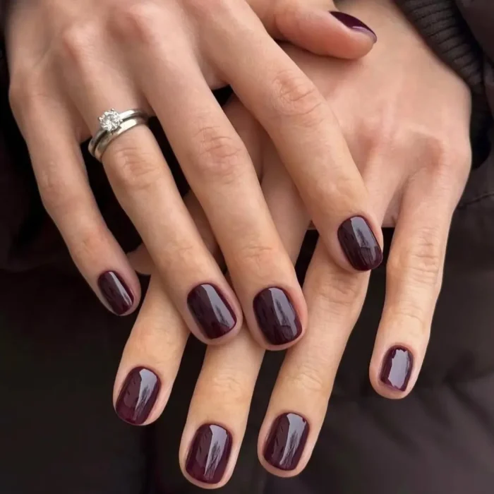 A pair of hands with a glossy, deep black cherry manicure on short, squared-off nails.