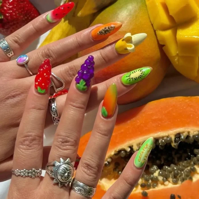 A pair of hands with long, colorful 3D fruit nail art, including sculptural banana, grape, and papaya designs.