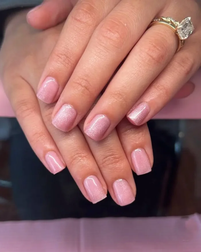 A pair of hands showing a soft baby pink cat eye manicure on short, neatly squared nails, creating a subtle radiant glow.