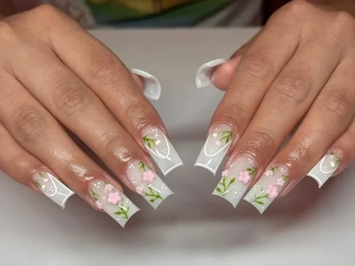 A close-up of cute white nail designs, featuring long square nails with a milky base, delicate pink flowers, and green leaf accents.