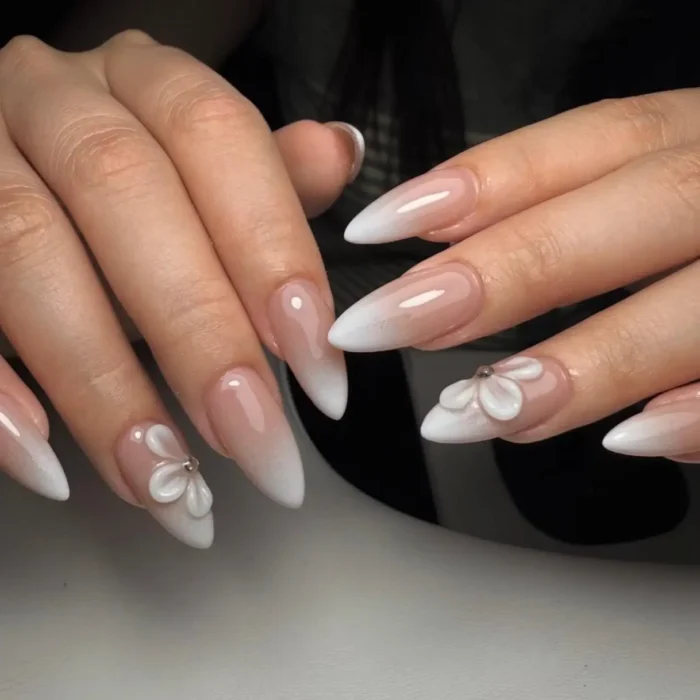 A close-up of cute white nail designs on long almond nails, featuring a white ombré or 'baby boomer' effect and elegant 3D flower accents.