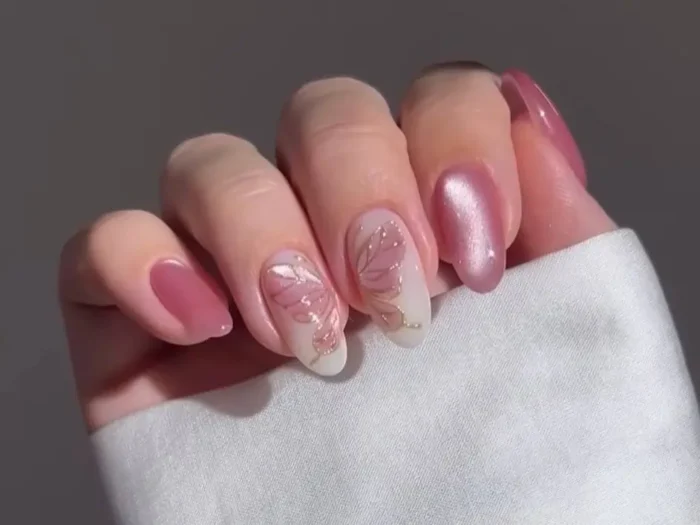 A close-up of a hand with a chic manicure featuring shimmery rose gold nails and accent nails with a gold-outlined butterfly wing design.