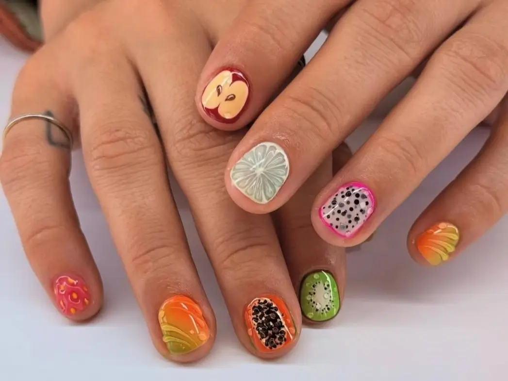 A pair of hands showcasing vibrant and realistic fruit nail designs on short nails, with a different fruit slice like apple, kiwi, and dragon fruit on each finger.