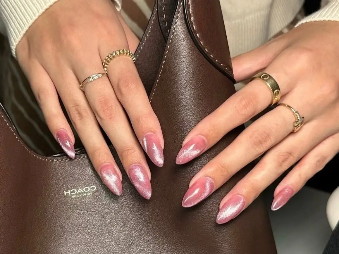 A close-up of a person's hands with a chic pink cat eye nail design on long stiletto nails, resting on a brown leather hand bag.