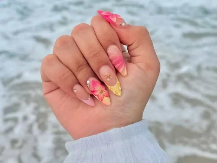 Summer 3D Flower Nails article thumbnail: Hand showcasing almond nails with tropical pink and yellow ombre, 3D flowers, and pearl accents by the ocean.