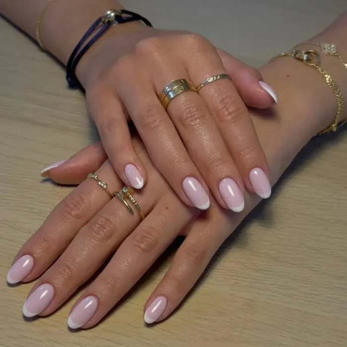A woman's hands with a classic French manicure on perfectly shaped short oval nails, adorned with gold rings.