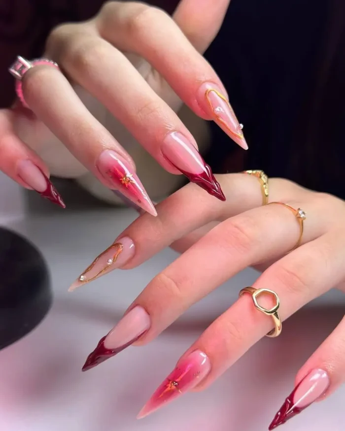 A close-up of long stiletto nails with fiery burgundy tips, gold lines, and pearl details.