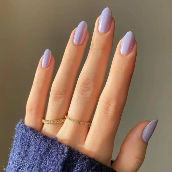 A hand with medium-length oval nails painted in a shimmering lavender polish, against a cozy blue sweater sleeve.