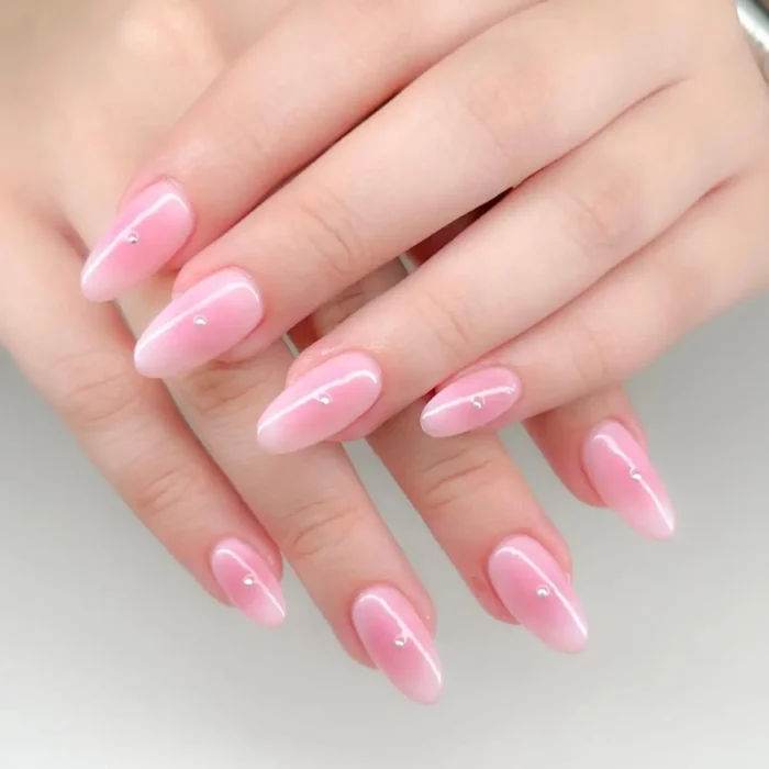 A close-up of almond-shaped nails with a pink and white aura ombre effect, each nail accented with a single small rhinestone.
