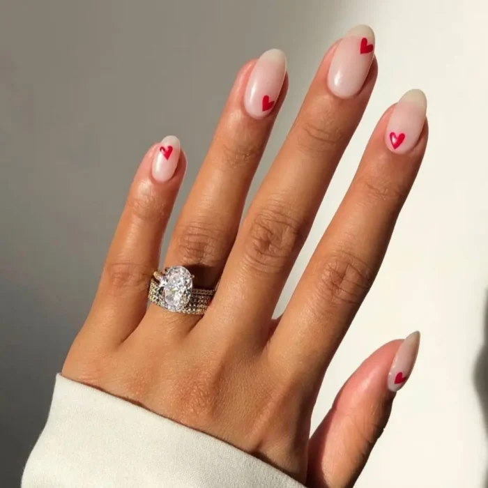 A close-up of a hand with a diamond ring, featuring a simple oval nail design with dainty red hearts on a sheer nude polish.