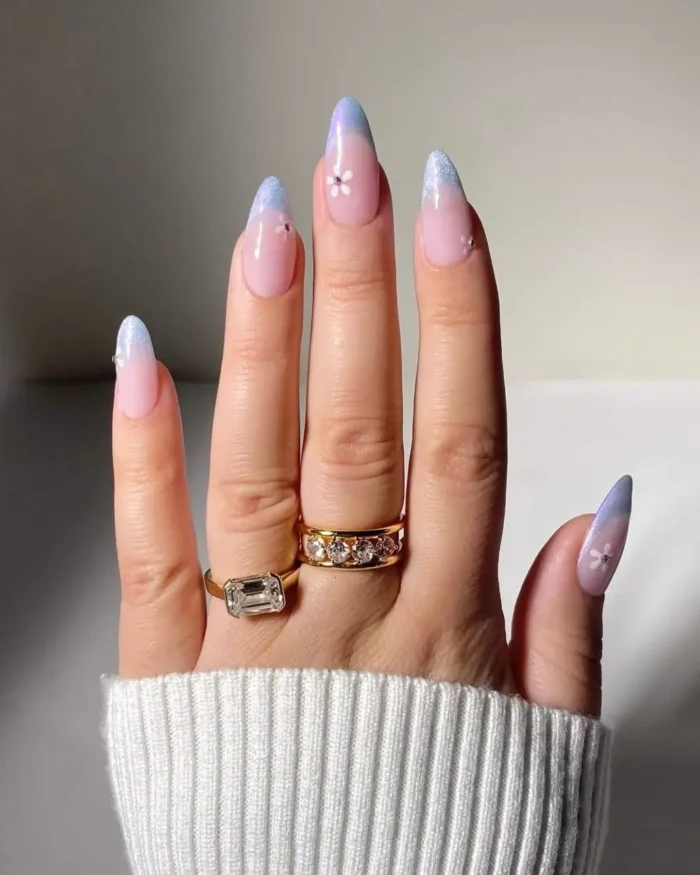 A close-up of long almond nails with a periwinkle gradient cat eye french tip and a small flower accent.