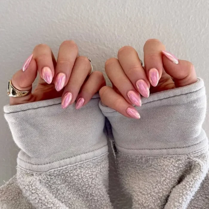 Hands with almond-shaped nails painted in a reflective, chrome-finish pink polish, held against a grey sweatshirt.