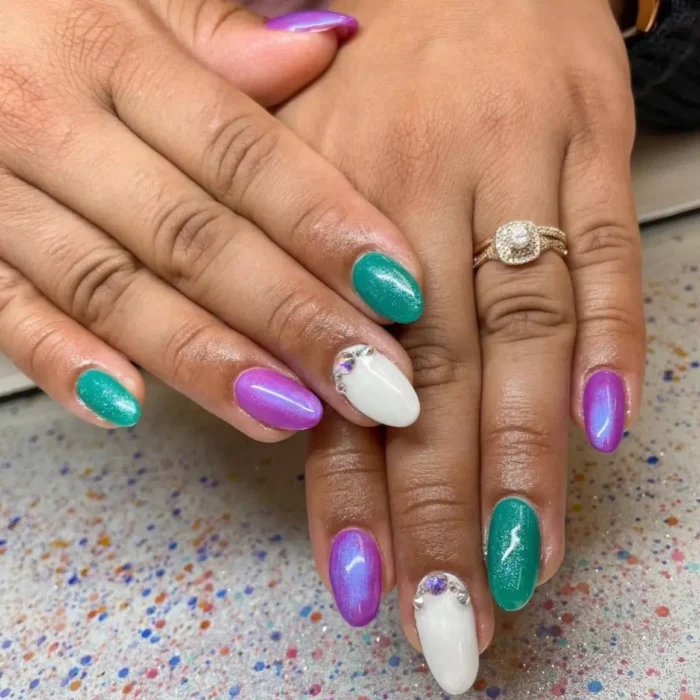 Hands showing a colorful oval nail design with purple, teal, and white polish, plus a simple rhinestone accent.