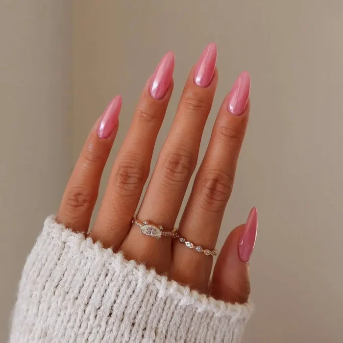 A hand with long, stiletto-shaped nails coated in a shimmery, pearlescent pink polish against a neutral background.