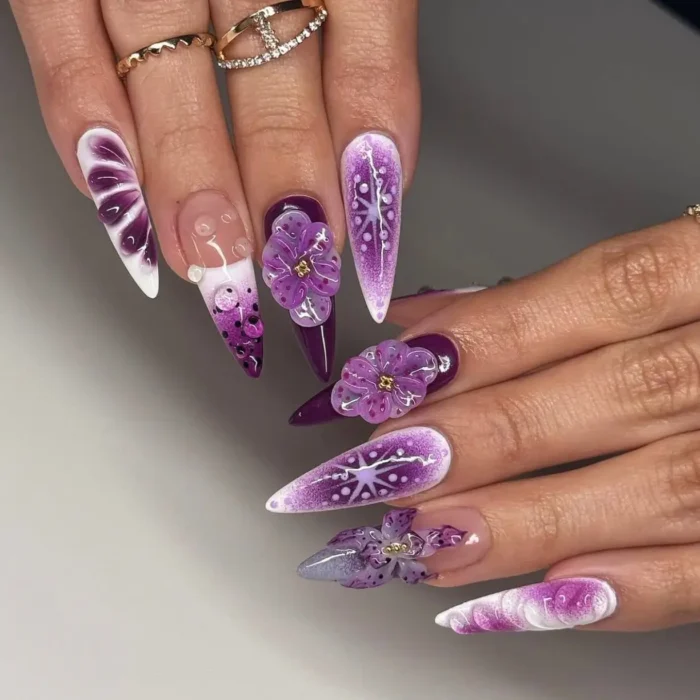 Long stiletto nails with various purple designs, including swirls, snowflakes, and 3D flowers with gold centers.
