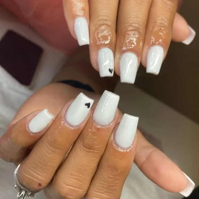 Hands showing a simple white nail art design with a tiny black heart accent on a square-shaped nail.