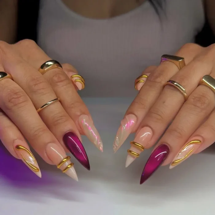 Long stiletto nails with iridescent pink, deep magenta cat-eye effect, and gold 3D rings and wavy line accents.