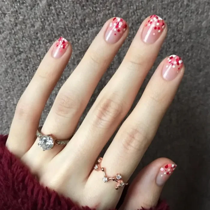 A hand with short, rounded nails on a sheer pink base, with a gradient of red and white confetti glitter at the tips.