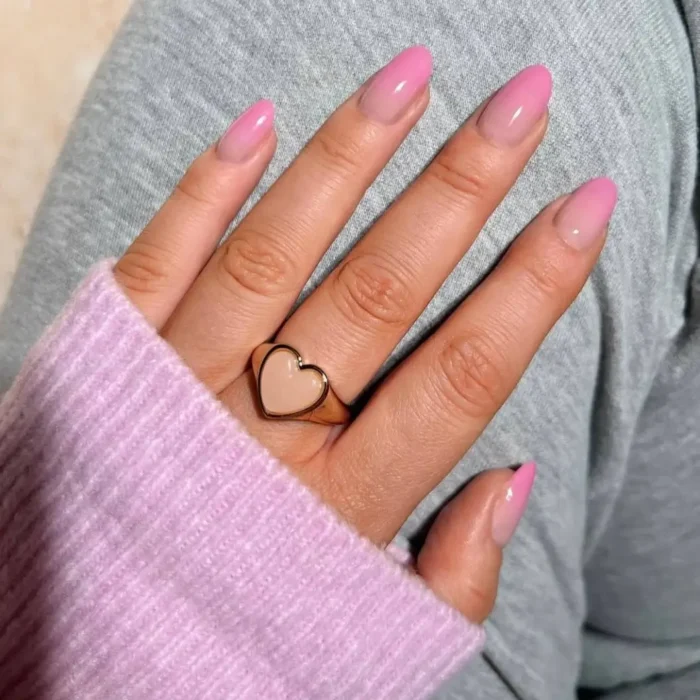 A hand wearing a pink sweater and heart ring, showcasing a pink ombré design on long oval nails.