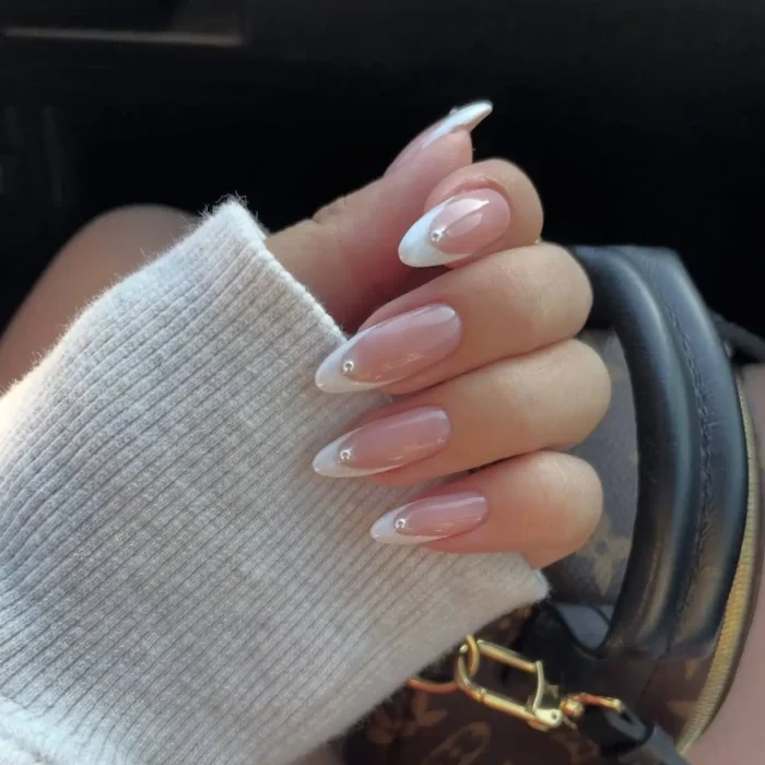 A hand with almond-shaped nails featuring a simple white nail idea of a French manicure with a single rhinestone accent.