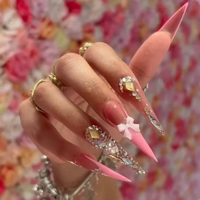 Extremely long pink stiletto nails with large crystal clusters and a white 3D bow.