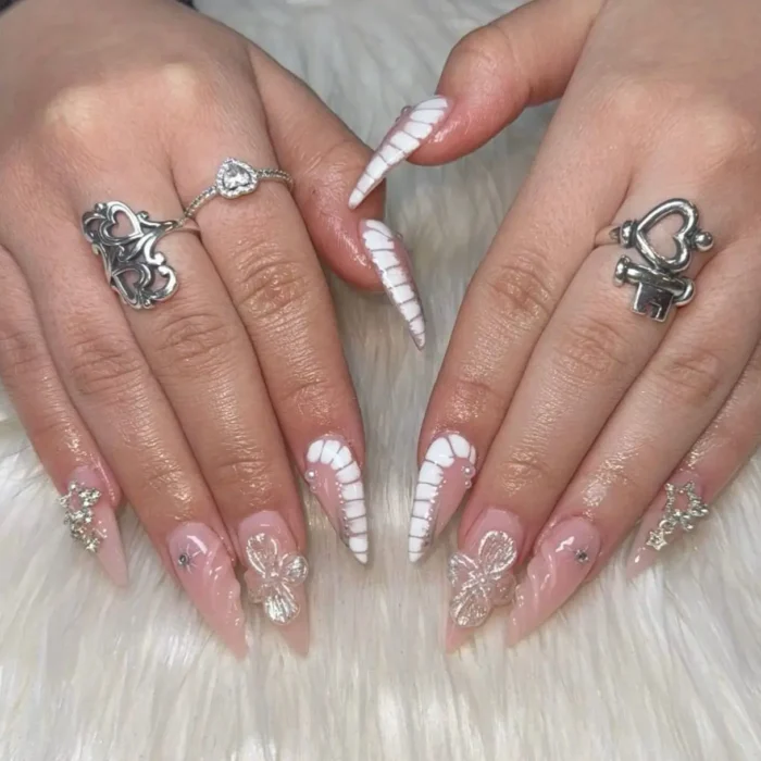 Light pink stiletto nails with white corset texture, 3D butterfly charms, and star rhinestones.