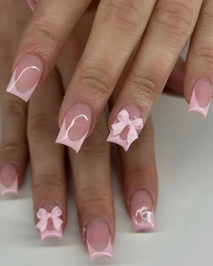 A close-up of a pink French manicure on square nails, with some nails featuring a raised 3D bow accent.