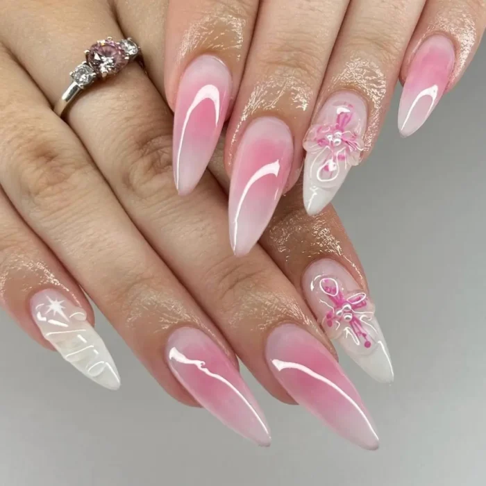 A close-up of long stiletto nails with a pink and white ombre design, featuring intricate 3D pink floral accents and white details.