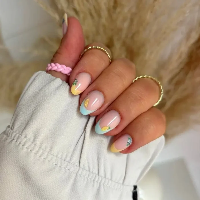 A hand showing a simple oval nail design with pastel yellow and blue French tips and tiny lemon nail art.