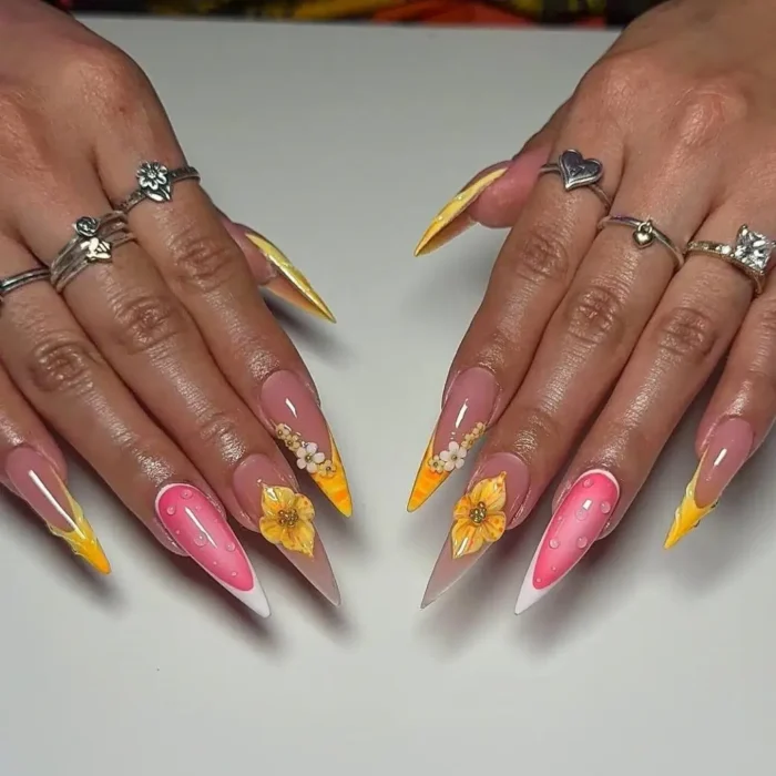 Long stiletto nails with yellow and orange gradient tips, some with 3D yellow flowers, and pink accent nails with white raised dots.