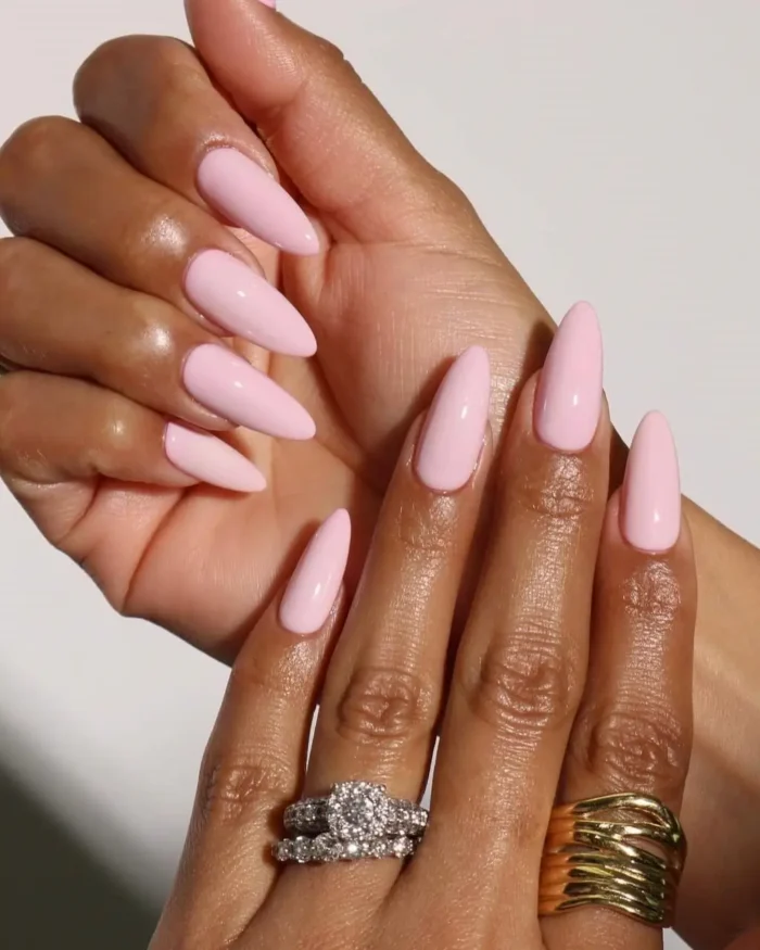 Hands with long, almond-shaped nails painted in a glossy, opaque light pink polish, adorned with rings.
