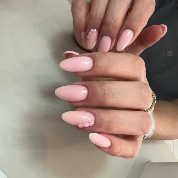 A hand showing a simple design on medium oval nails, featuring a soft pink polish with delicate 3D floral accents.