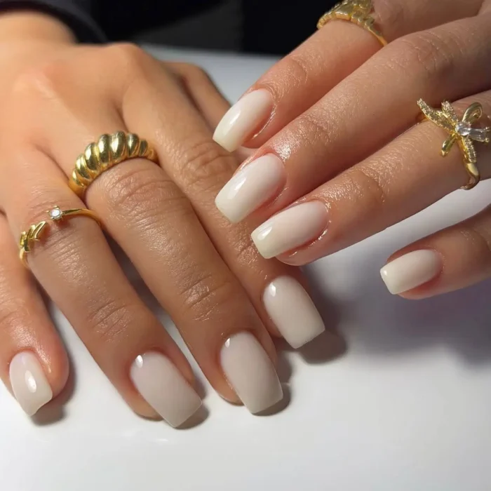 A pair of hands with gold rings showing a simple white nail design in a sheer, milky finish on square nails.