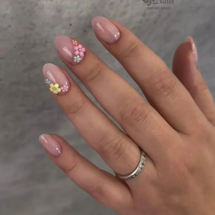 A hand with a simple oval nail design featuring a sheer pink base and delicate 3D pastel flower accents.