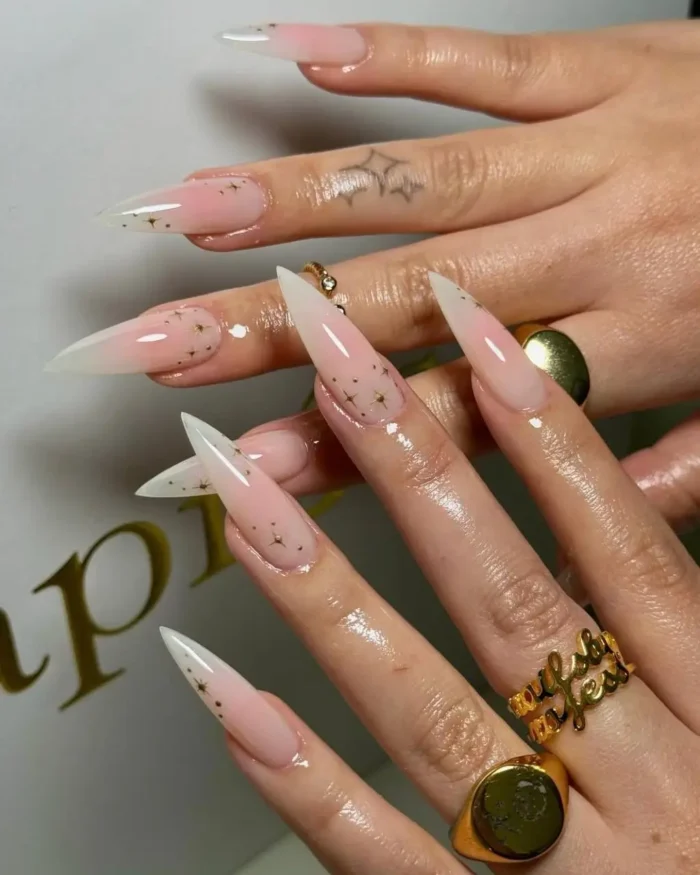 Long stiletto nails with a pink to white ombre, decorated with small gold star glitter.