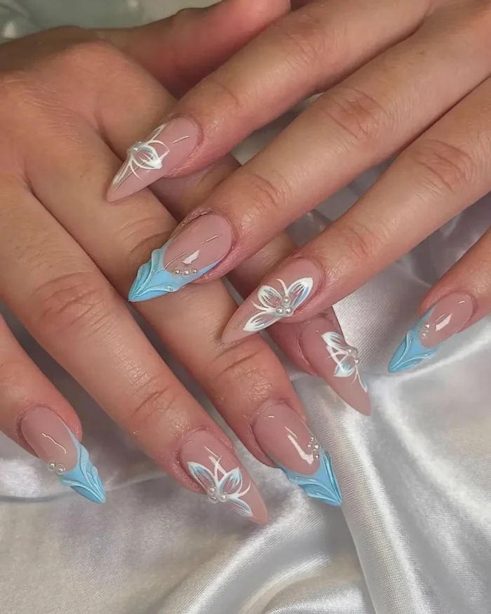 Medium stiletto nails with light blue and white tips, intricate white floral designs, and small pearl embellishments.