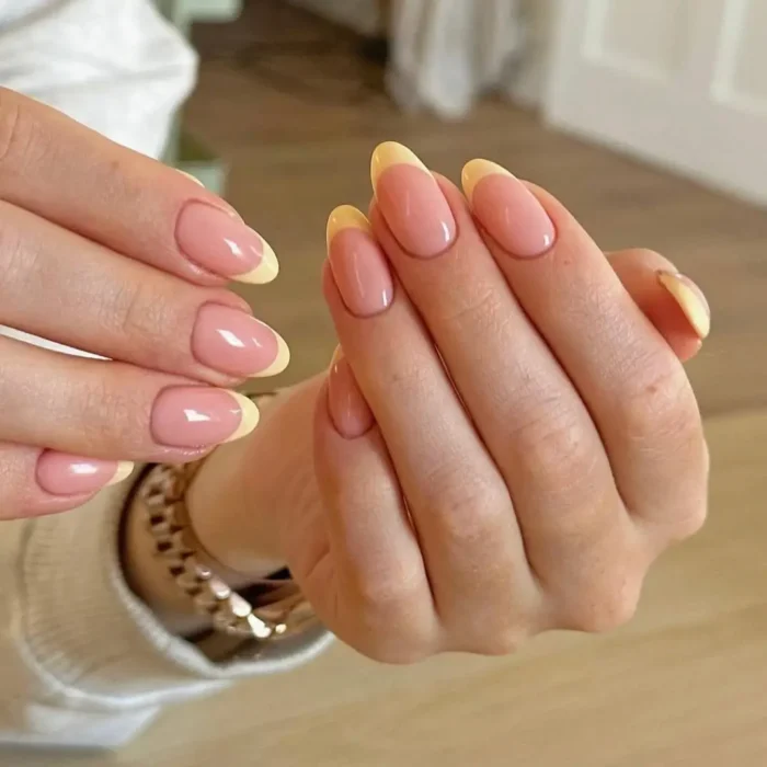 A pair of hands with a simple oval nail design featuring a French manicure with buttery yellow tips.