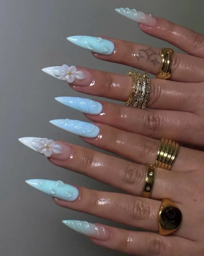 Long stiletto nails in light blue with white tips, featuring 3D white flowers, raised water droplets, and textured wavy designs.