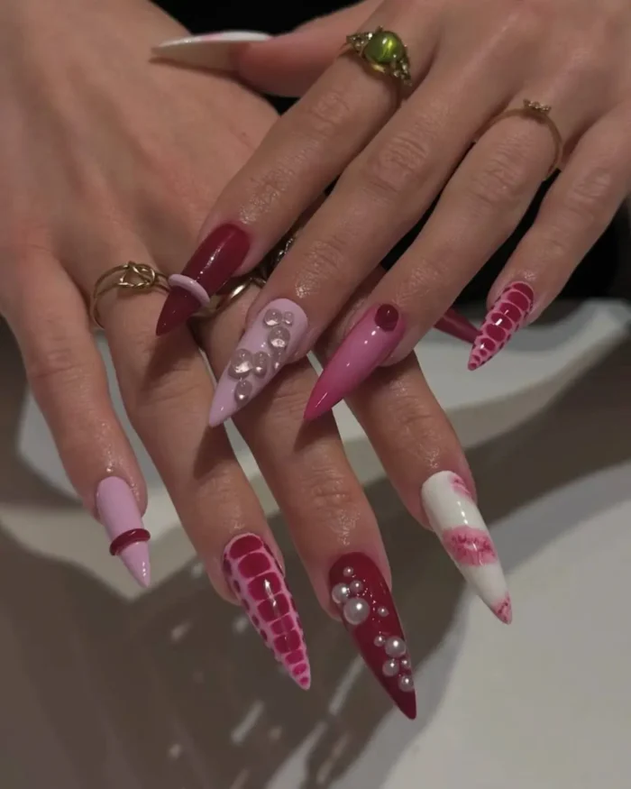 Long stiletto nails in shades of pink and berry red, featuring striped textures, pearl clusters, and ring embellishments.