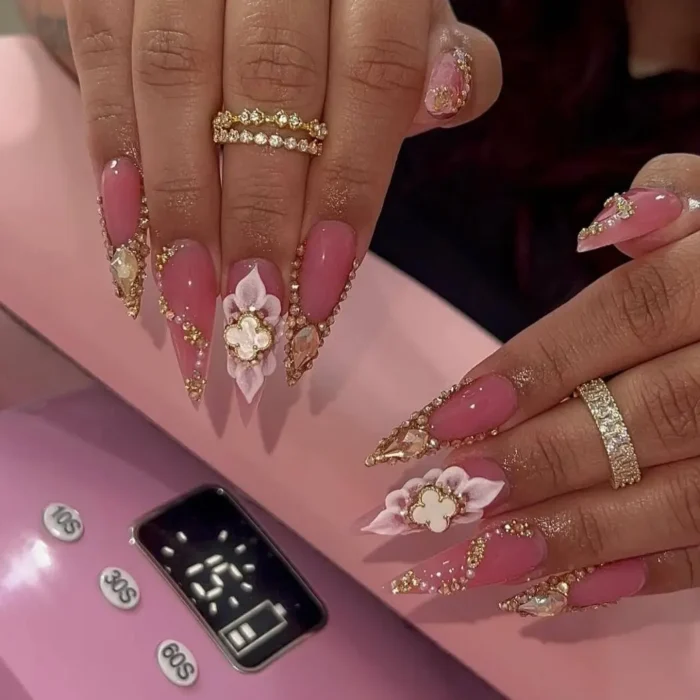 Glamorous pink stiletto nails with oversized gems, 3D flowers, and gold rhinestones.