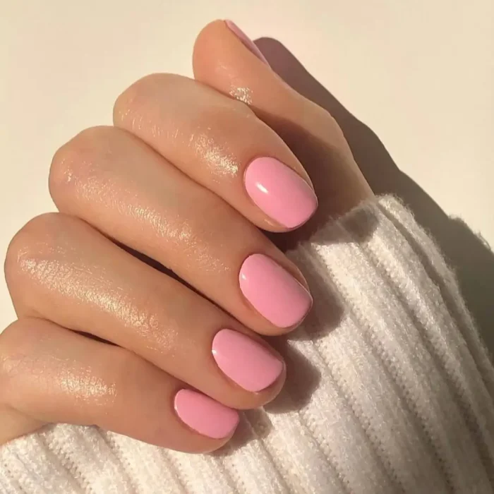 A hand with short, round nails painted in a glossy bubblegum pink polish, resting on a white knit sweater.