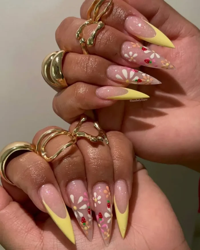 Long stiletto nails with yellow French tips, featuring hand-painted white and pink daisies and small red strawberries.