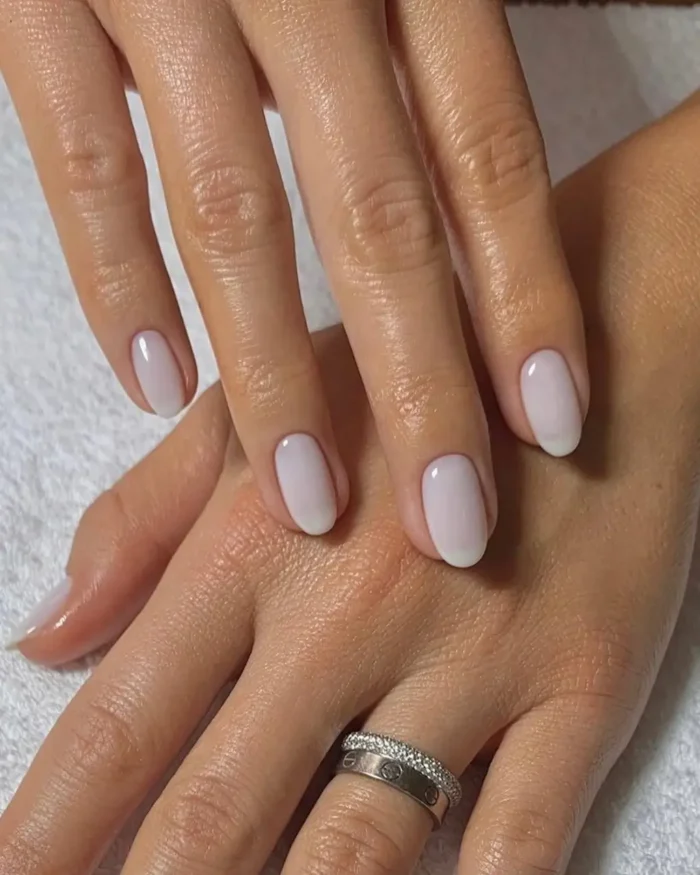 A close-up of hands showcasing a beautiful, simple milky white manicure on short oval nails.