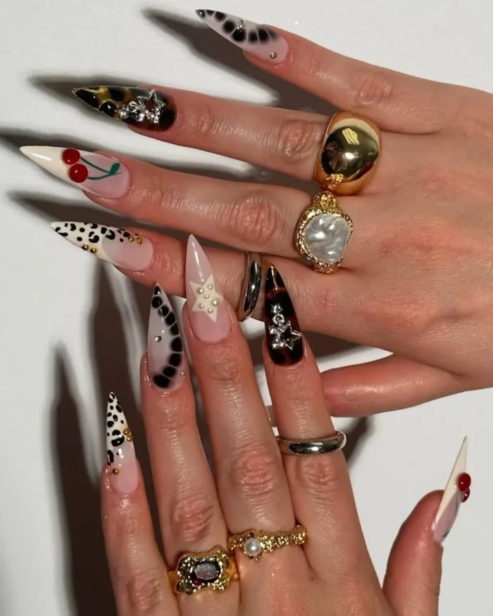 Long stiletto nails with various designs including leopard print, tortoiseshell, cherries, and accents of studs, pearls, and stars.