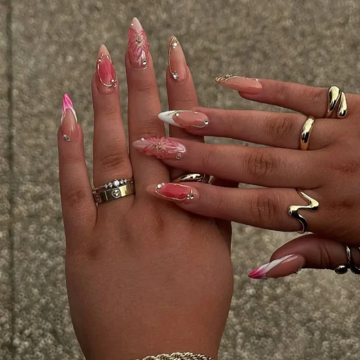 Medium stiletto nails with pink bases and some pink French tips, decorated with 3D pink flowers, rhinestones, and gold lines.